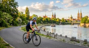 A professional editorial photograph of a cyclist riding along a scenic, sun-drenched riverside bike